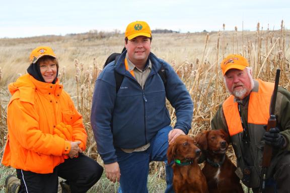 Three people standing in a grassy field, with one person holding a hunting dog