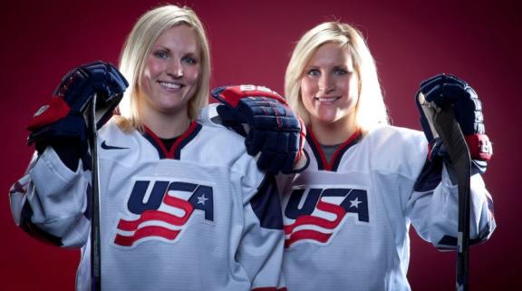 Lamoureux Twins pose with their sticks, smiling against a red background