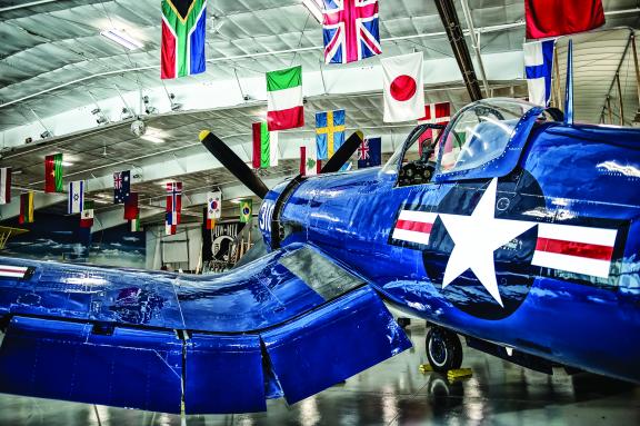 A restored blue military aircraft is on display inside a museum, with various national flags hanging from the ceiling