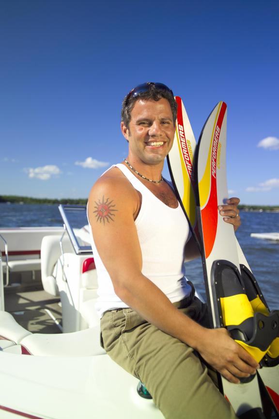 A person in a tank top and khaki pants holds a colorful wakeboard while smiling, standing on a boat 