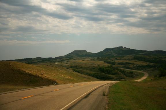 A winding road stretches into a hilly, green landscape under a cloudy sky, with distant hills and mountains visible
