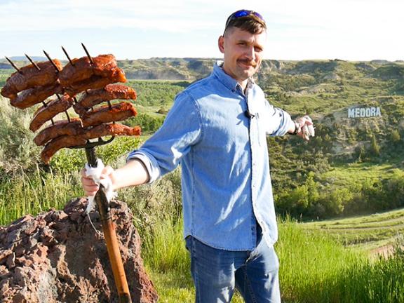 Lyubomyr Shkandriy is smiling and holding a large skewer with multiple sausages in a scenic area with green hills