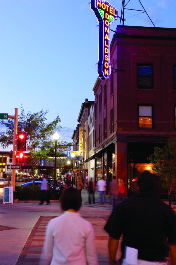 The exterior of a downtown area at dusk, featuring a brightly lit theater marquee and a lively street scene with people walking and gathering