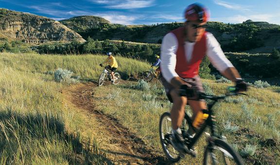 Two mountain bikers ride on a dirt trail through a rugged landscape with hills and sparse vegetation
