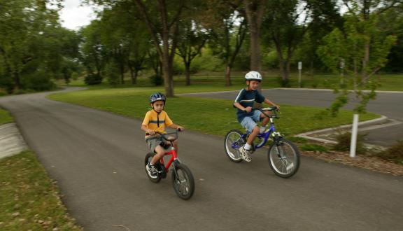 Two people are cycling on a paved trail in a park