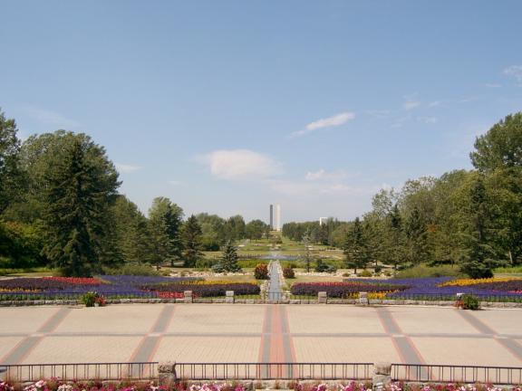 A spacious outdoor area with a tiled floor and neatly arranged flower beds, leading towards a fountain and a garden in the distance