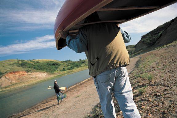 A close-up of a person carrying a canoe over their shoulders while walking along a riverbank, with another person