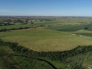 aerial view of fort clark state historic site in north dakota, showing circular earth lodge depressions, open grasslands, winding paths, nearby farmland, and the missouri river valley landscape. photo